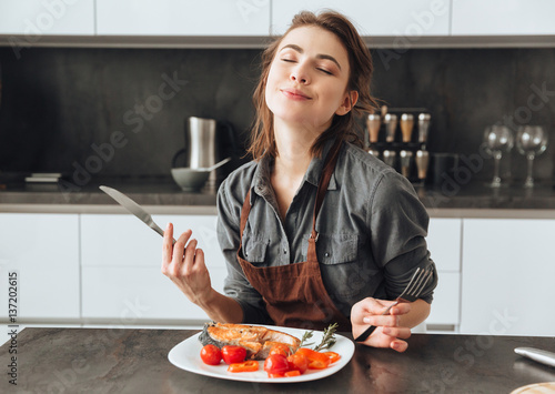 Valokuva Woman sitting in kitchen while eating fish and tomatoes.