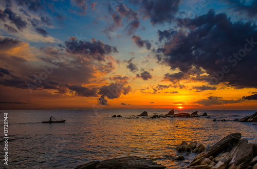 Beautiful sunrise over lake Malawi with rock formation and a canoe gliding trough the water under colorful cloudy sky. The bright orange sunlight on the horizon gradually changes color to blue