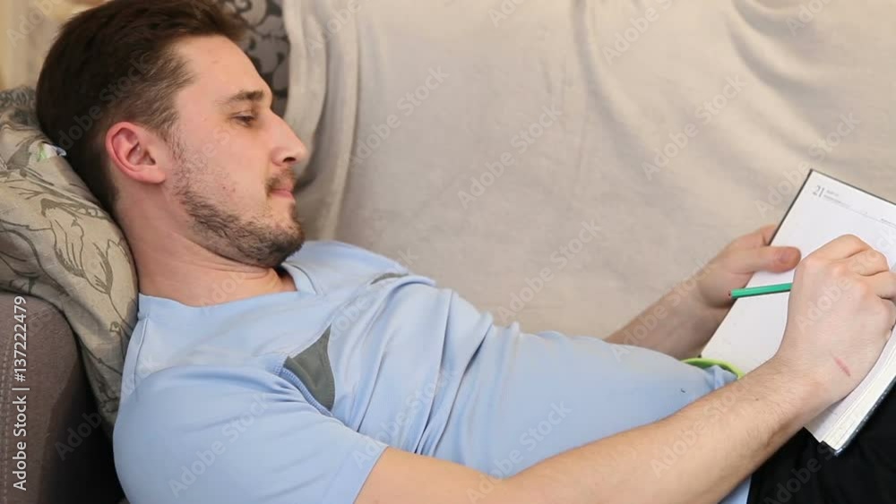 man takes notes in a notebook lying on the couch