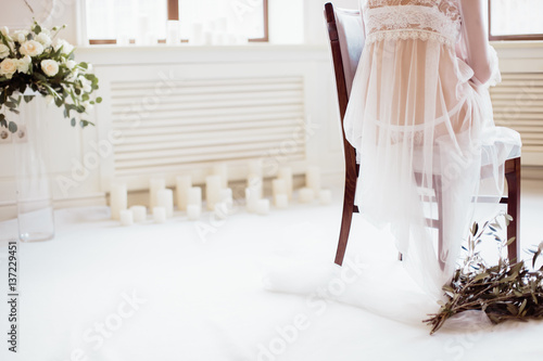 girl in white transparent boudoir gown sitting on a chair with his back on the background of beautiful scenery with flowers and window