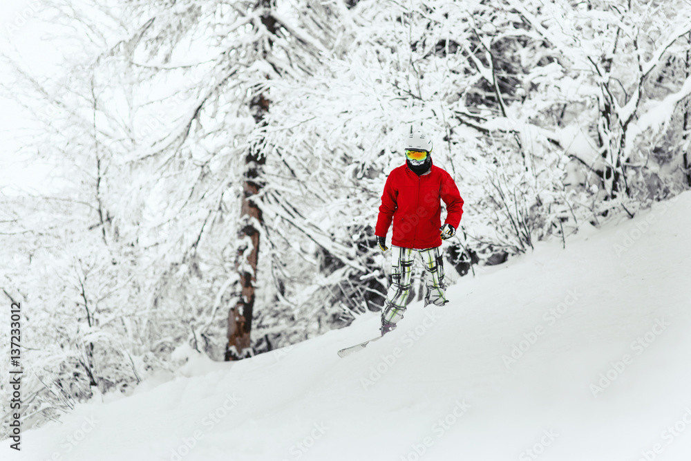 Man in red ski jacket and white helmet goes down the snowed hill in the forest