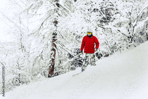 Man in red ski jacket and white helmet goes down the snowed hill in the forest