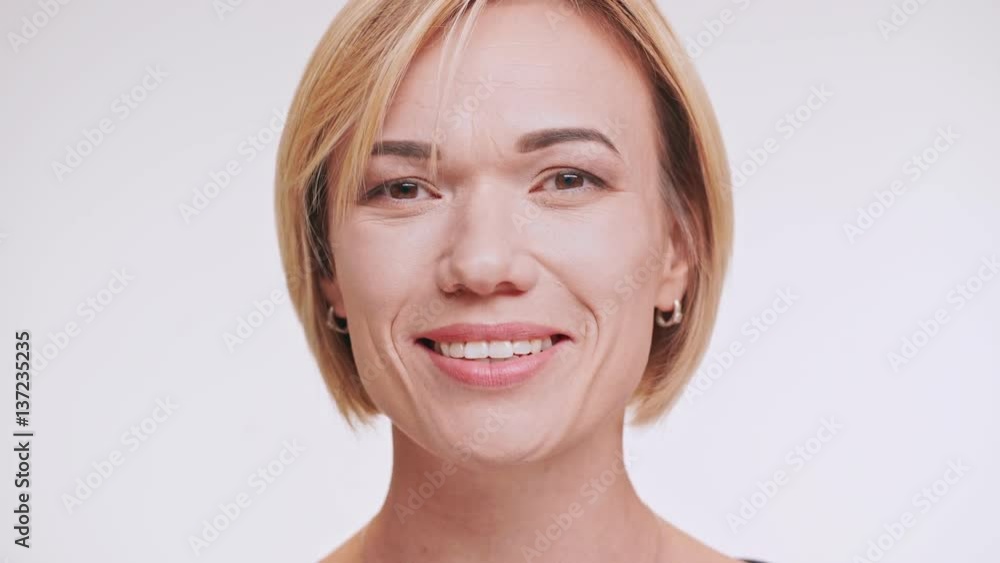 Blonde Caucasian middle-aged female smiling at camera winking eye showing ok and touching hair on white background. Close up footage Blonde Caucasian middle-aged female smiling at camera winking eye showing ok and touching hair on white background. Close up footage