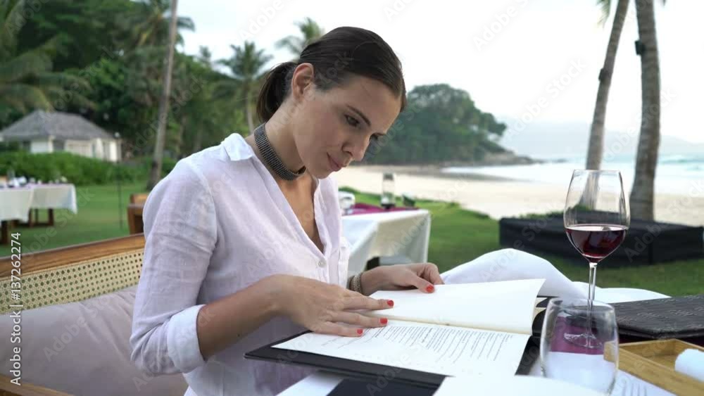 Young, elegant woman reading menu sitting by table in beach restaurant
Young, elegant woman reading menu sitting by table in beach restaurant