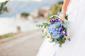 Bride holds wedding bouquet from blue flowers hydrangea and stands on light background of sea on way to wedding ceremony. Bride is dressed in white lace wedding dress.