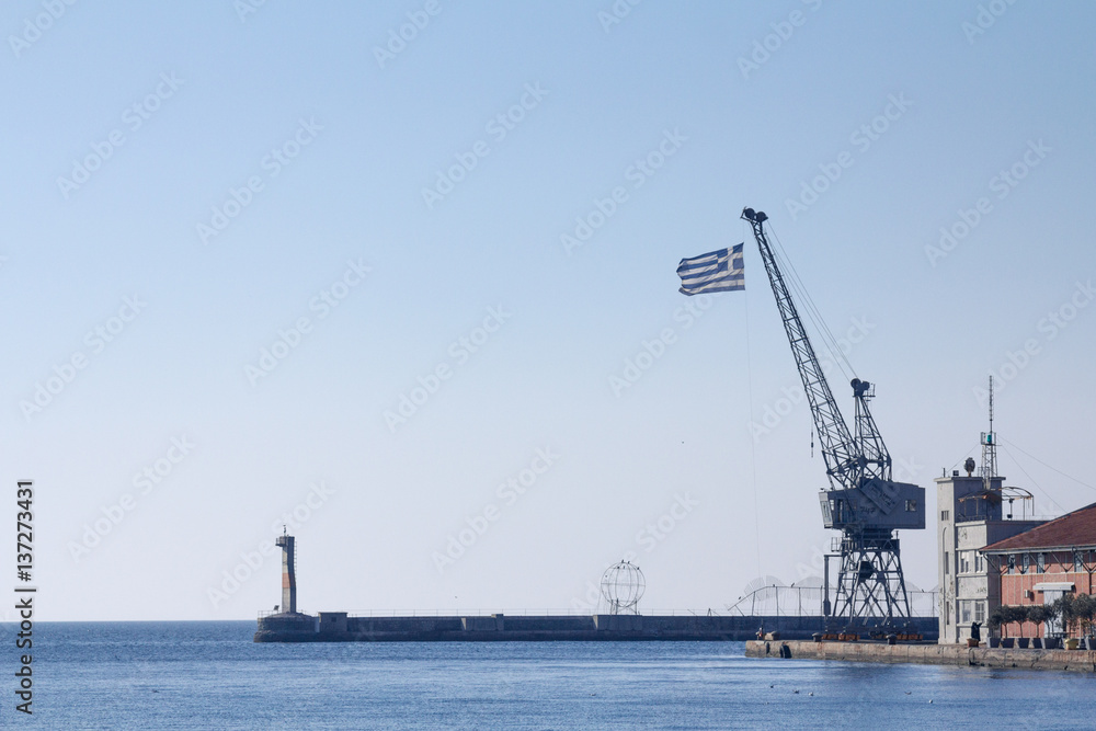 Fototapeta premium Greek flag on a crane on Thessaloniki pier, Greece..Picture of a Greek flag on a crane seen in Thessaloniki, Greece, on the iconic pier of the old port