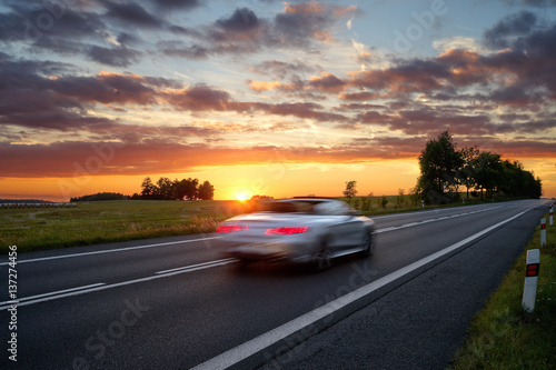 Speeding motion blur white car on the road in a rural landscape at sunset.