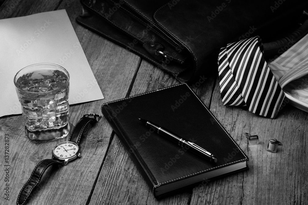 business accessories - notebook, pen, rich portfolio, watch, tie, glass of  whiskey with ice on wooden background, black and white Stock Photo