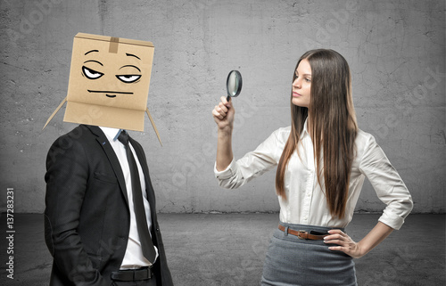 Tableau sur toile Woman is looking through a magnifier at a businessman with a box on his head wit