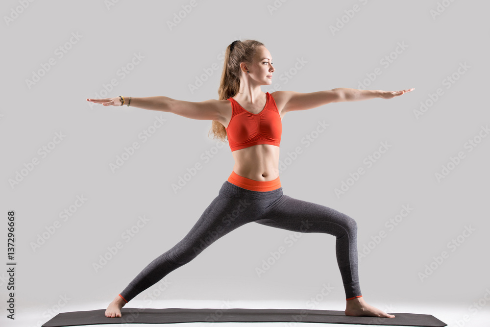 Young attractive yogi woman practicing yoga, standing in Warrior Two exercise, Virabhadrasana II pose, working out, wearing sportswear, red sports bra, pants, indoor full length, isolated, grey studio