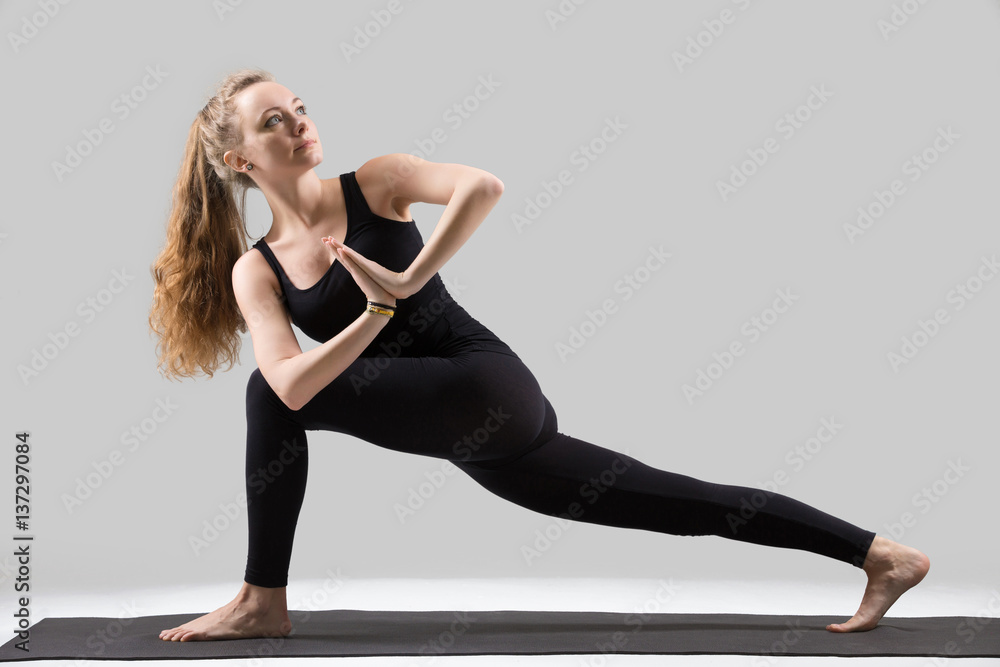 Young attractive yogi woman practicing yoga, standing in Revolved Side Angle exercise, Parivrtta Parsvakonasana pose, working out, wearing sportswear, black tank top, pants, full length, grey studio