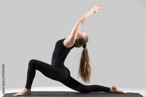 Young attractive yogi woman practicing yoga, stretching in Horse rider exercise, anjaneyasana pose, working out, wearing sportswear, black tank top, pants, indoor full length, isolated, grey studio photo