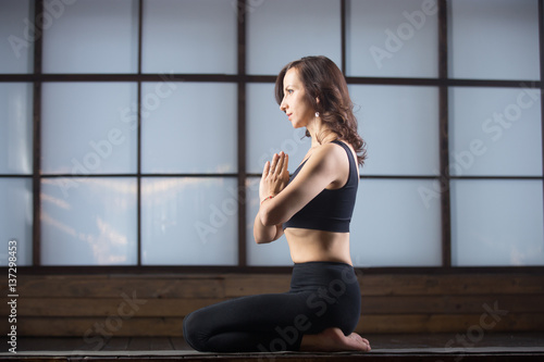 Young attractive woman practicing yoga, sitting in vajrasana exercise, seiza pose with namaste, working out, wearing sportswear, black tank top, pants, indoor full length, studio evening practice