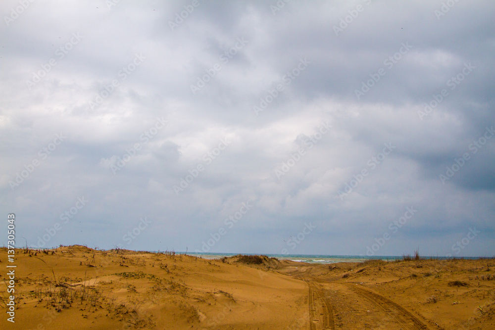 Photo of a sandy road to beach.  There is only sand until the sea. Tyre trails are also visible. Most of the photo is covered with clouds.