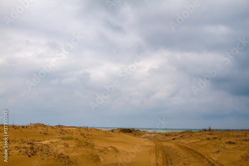 Photo of a sandy road to beach. There is only sand until the sea. Tyre trails are also visible. Most of the photo is covered with clouds.
