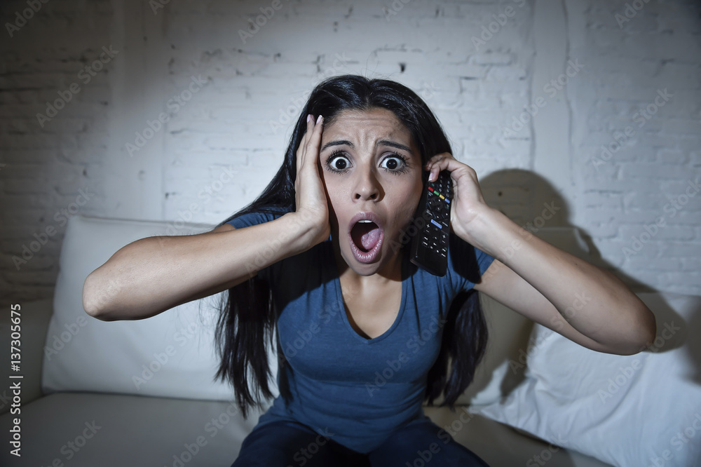 Naklejka premium latin woman at home sofa couch in living room watching television scary horror movie or suspense thriller