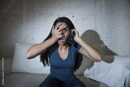 latin woman at home sofa couch in living room watching television covering eyes horrified