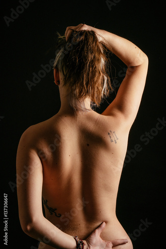 Chica mostrando  la espalda con tatuajes y con los brazos levantados. Foto de estudio con fondo negro.