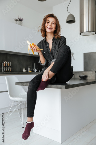 Happy woman sitting at kitchen eating banana indoors фототапет