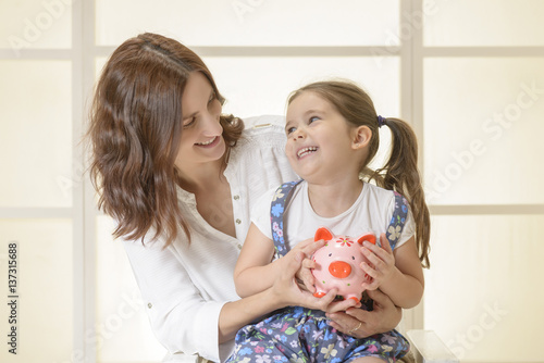 Happy mother giving piggy bank to her beautiful little daughter. Family budget and savings concept. Education, school and money saving concept. Junior Savings Account concept
