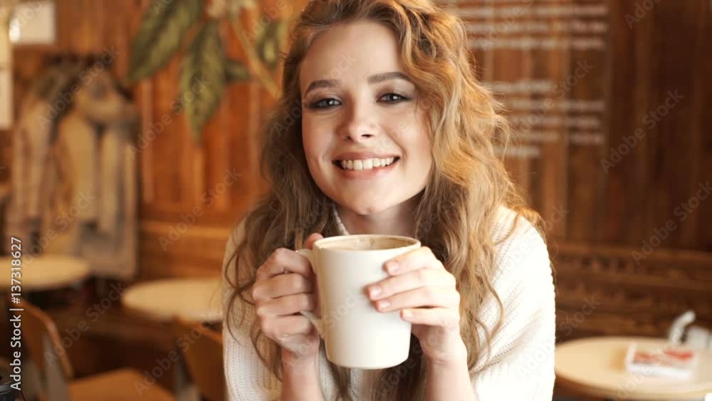 Young attractive woman smiling to camera in cafe. Happy girl drinking coffee and relaxing in cafe, smiling and looking at camera. Beautiful woman with cup of coffee sitting in the coffee shop Young attractive woman smiling to camera in cafe. Happy girl drinking coffee and relaxing in cafe, smiling and looking at camera. Beautiful woman with cup of coffee sitting in the coffee shop