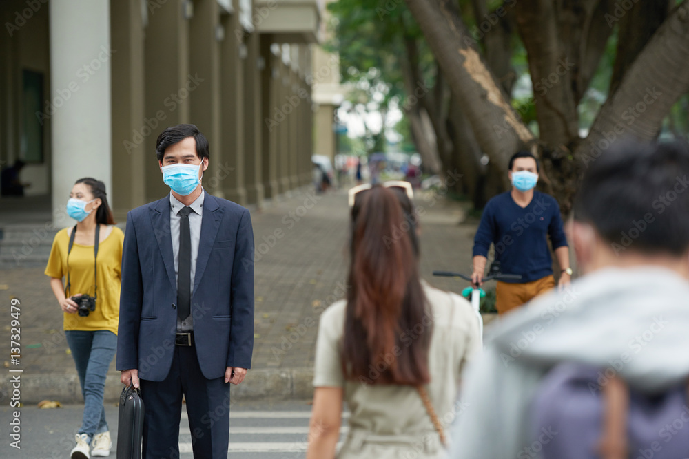 Portrait of middle-aged Asian businessman wearing facial mask in order to protect himself from smog while crossing road with other people