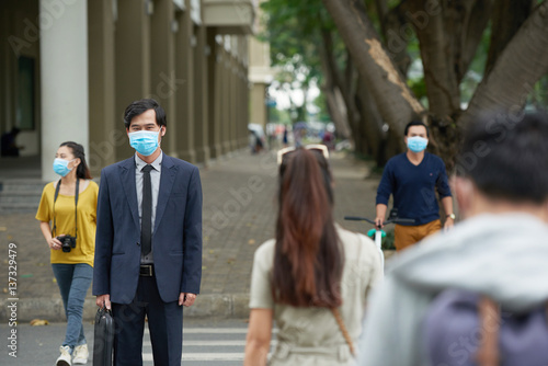 Portrait of middle-aged Asian businessman wearing facial mask in order to protect himself from smog while crossing road with other people