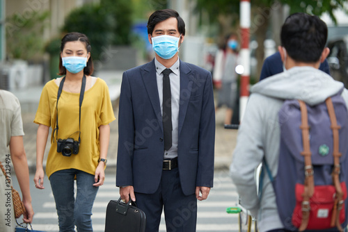 Asian businessman in protective mask looking at camera seriously while standing against pedestrian crossing