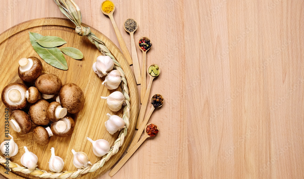 Braid of garlic, mushrooms and bay leaves on a round cutting board light wooden table. Nearby are several small wooden spoons with different spices. Top view. Braid of garlic, mushrooms and bay leaves on a round cutting board light wooden table. Nearby are several small wooden spoons with different spices. Top view.
