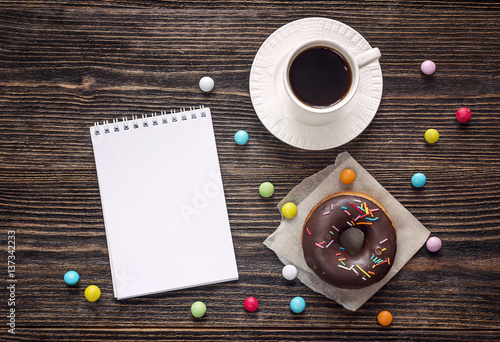 Open blank notebook, cup of coffee and a chocolate donut on a wooden table. Space for text.