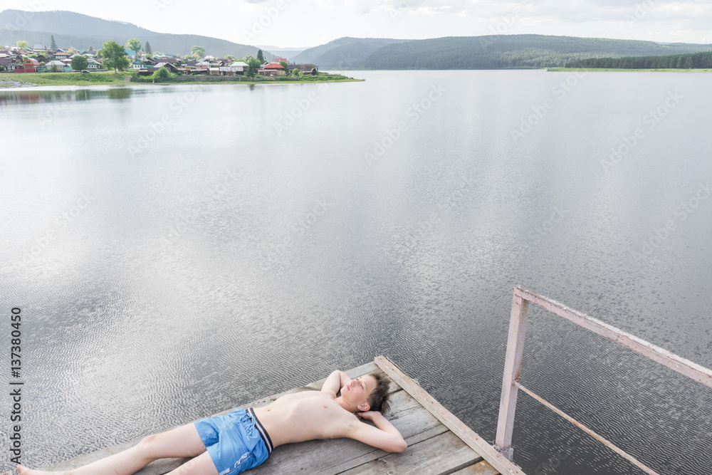 Fototapeta premium Young boy relaxes on a tower for jumping into the water. Boy resting near the lake. Swimmer in shorts. summer rest. teenager sunbathe on the waterfront 