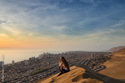 Young woman sitting at the sand dune and overlooking desert, city and ocean