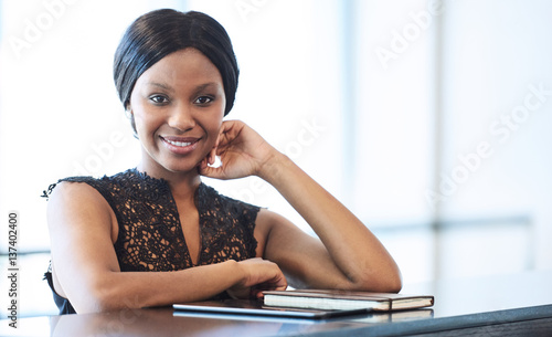 Successful black businesswoman looking into camera while seated at counter with bright backlighting from large windows behind her, and her notebook and electronic tablet in front of her.