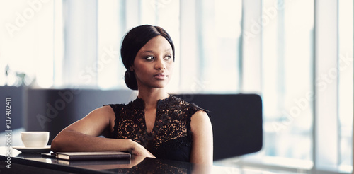 фотографія Beautiful young black woman wearing a stunning black dress while seated at a cafe counter with her coffee, notebook and tablet laying in front of her on the counter top