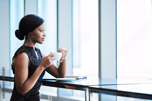 Elegant African American woman thoughtfully looking out the large window while holding her cup of tea in her hands, resting one elbow on the counter maintaining regal posture photo