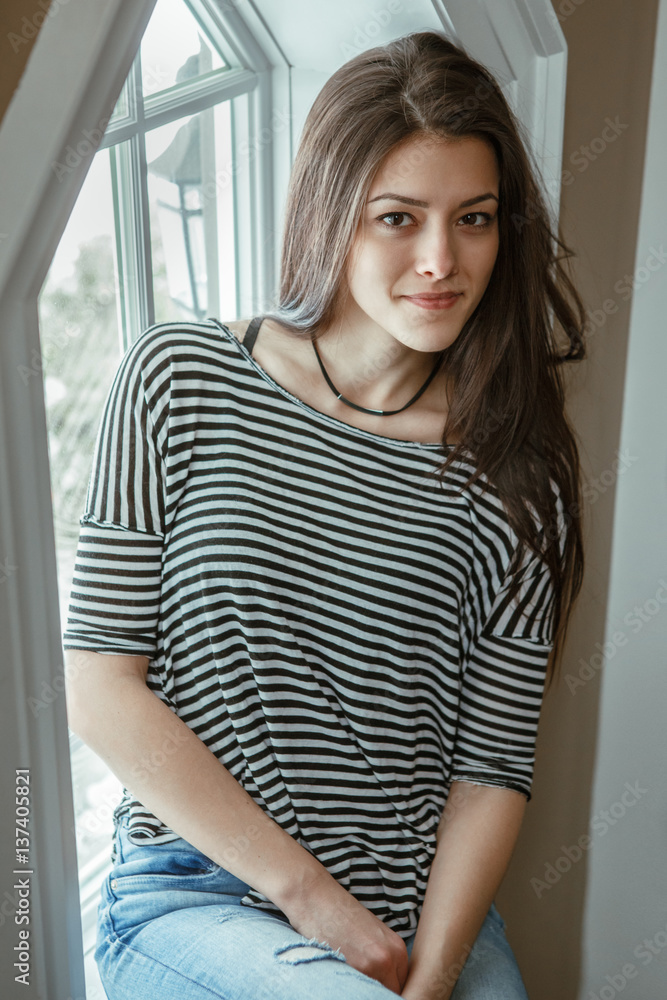 Closeup portrait of smiling Caucasian young beautiful woman model with messy long hair in ripped blue jeans and striped t-shirt sitting indoor by window, looking in camera, toned with filters.