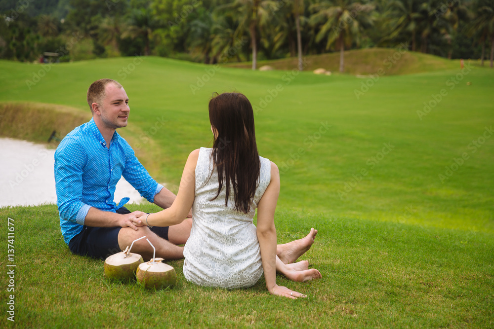 Young beautiful couple with coconuts holding their hands at golf course in summer. man wear the blue shirt and the girl in a white dress. Concept of honeymoon Young beautiful couple with coconuts holding their hands at golf course in summer. man wear the blue shirt and the girl in a white dress. Concept of honeymoon