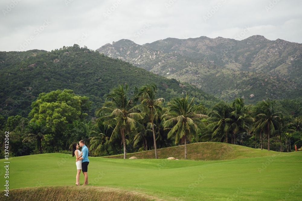 Young beautiful couple in love holding hands and walking at golf course in summer. man wear the blue shirt and the girl in a white dress. Concept of honeymoon Young beautiful couple in love holding hands and walking at golf course in summer. man wear the blue shirt and the girl in a white dress. Concept of honeymoon