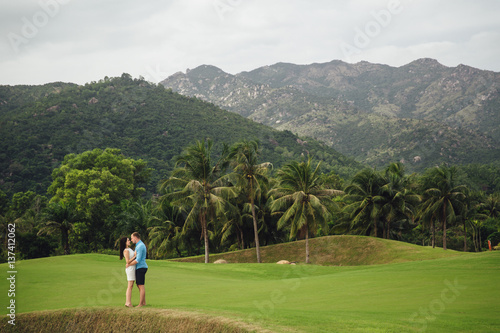 Young beautiful couple in love holding hands and walking at golf course in summer. man wear the blue shirt and the girl in a white dress. Concept of honeymoon