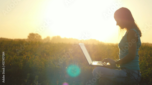 Young pretty woman typing on a laptop outdoors at beautiful sunset sitting on the grass with amazing lense flare effects