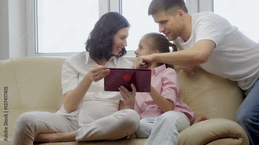 Parents showing digital tablet to their daughter at home.
Parents showing digital tablet to their daughter at home.
