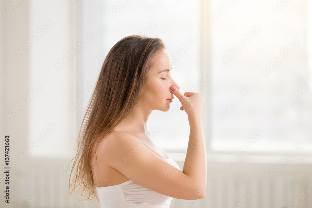 Side view portrait of young attractive yogi woman practicing yoga, making Alternate Nostril Breathing exercise, working out using nadi shodhana pranayama, white color room background, eyes closed Side view portrait of young attractive yogi woman practicing yoga, making Alternate Nostril Breathing exercise, working out using nadi shodhana pranayama, white color room background, eyes closed