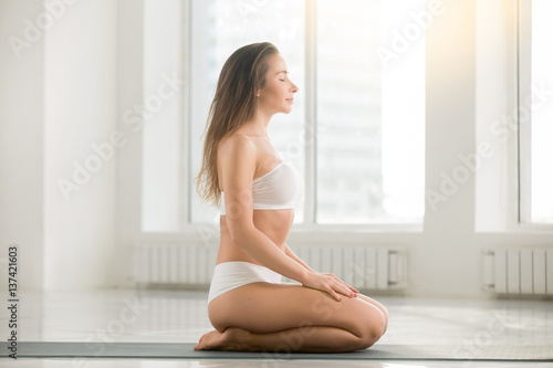 Young happy attractive yogi woman practicing yoga, sitting in vajrasana exercise, seiza pose, working out, wearing sportswear, sport bra, shorts, indoor full length, white color room background