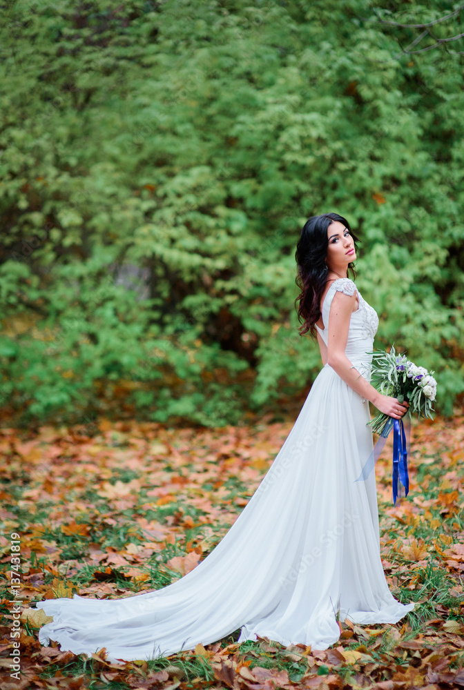 Stunning bride in dress with silver embroidery stands on fallen autumn leaves