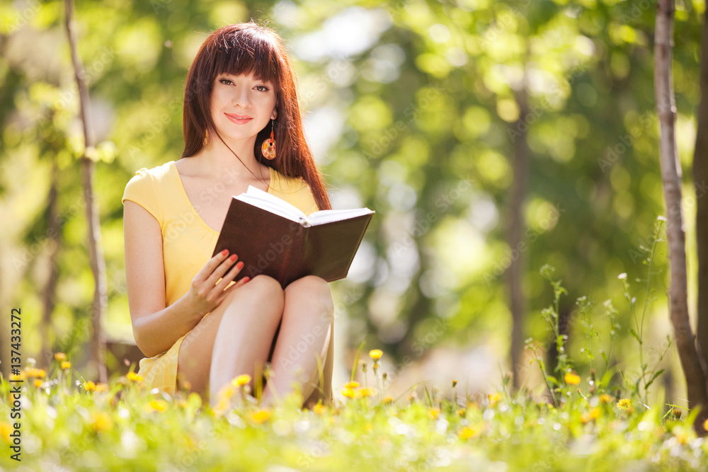 Young woman reading a book in the park with flowers. Beauty nature scene with colorful background, trees and flowers at summer season. Outdoor lifestyle. Happy smiling woman relax on green grass