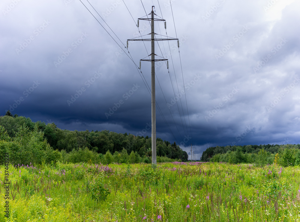 Electric pole stands in a green field. Over the field of storm clouds.