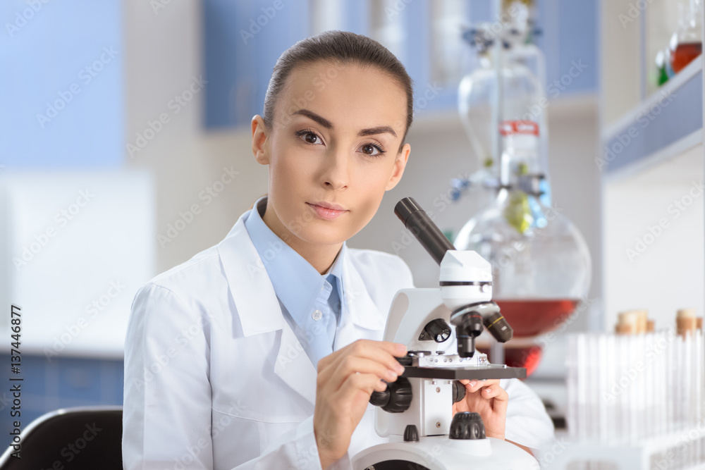 Young woman scientist in lab coat working with microscope and looking at camera Young woman scientist in lab coat working with microscope and looking at camera