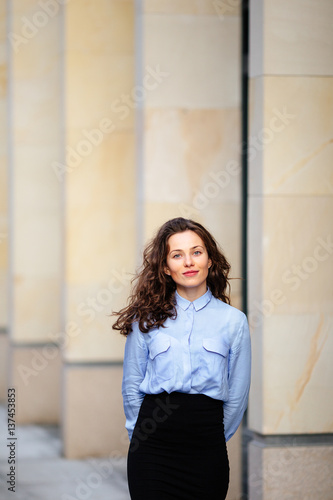 Free Download Brunette smiling woman standing near the wall of building, looking forward into camera - GFXLoad