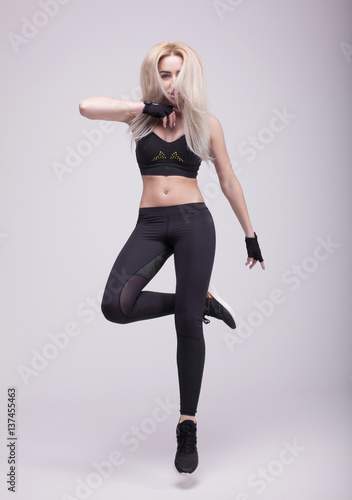 Beautiful young blonde fit woman with long hair, happily jumping on a grey background. Slim woman in sport clothes.
