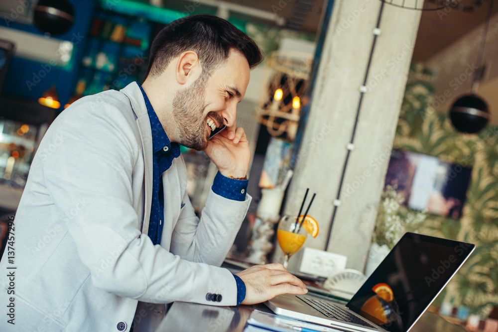 Fototapeta premium Young businessman talking on the phone and looking at laptop on table in cafe